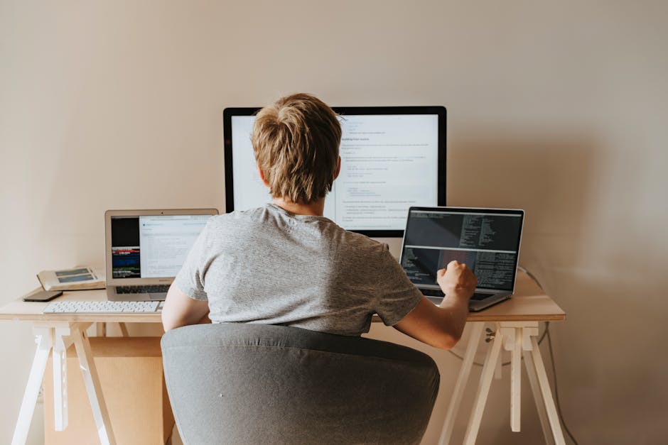A young man with short, light brown hair, wearing a light grey t-shirt, is seated at a wooden desk in a room with plain beige walls. He is facing away from the camera and working on a desktop computer
