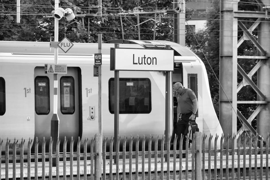 A man carrying a small black travel bag standing on the platform at Luton railway station with a modern train stopped nearby. The train has multiple windows with signs indicating first-class carriage 
